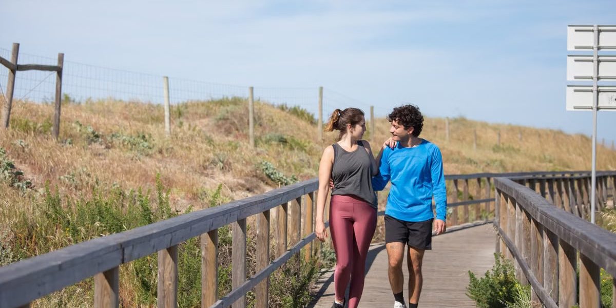 A couple in activewear strolling together on a sunny boardwalk in a natural setting.